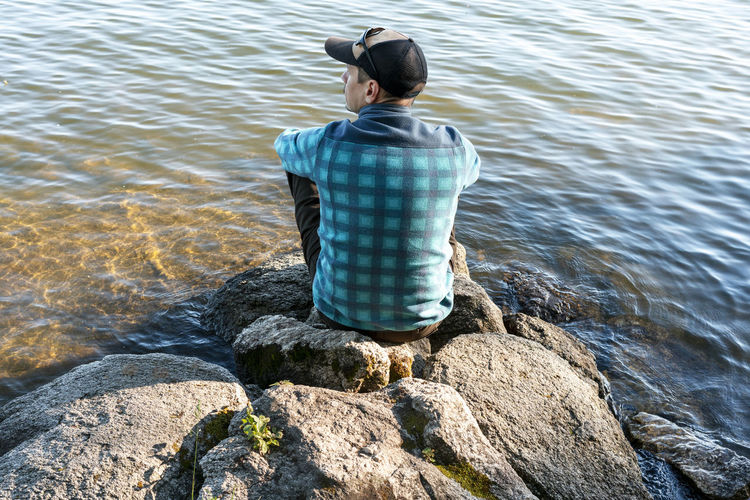 Young man in a cap looking at the view of the lake or river sitting on the stones enjoying nature 