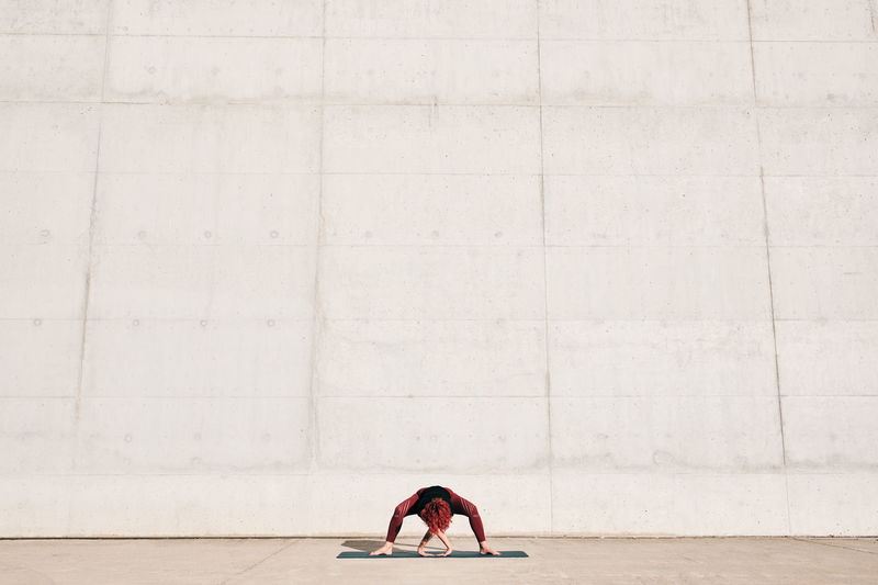 Unrecognizable fit female athlete in sportswear doing wide legged forward bend yoga pose on sports mat while training alone on street against concrete wall in sunny day