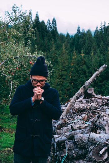 Man praying while standing against logs in forest
