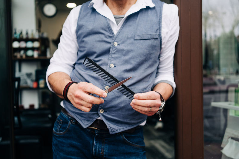 Crop unrecognizable male hairdresser in smart casual apparel with professional grooming tools at entrance door of beauty salon in town