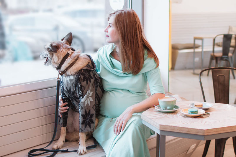 Young pregnant women is sitting in cafe with her dog in sweater and looking into the window.