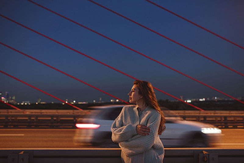 Thoughtful woman standing with arms crossed on bridge in city at night
