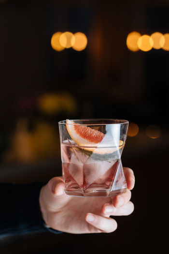 Cropped unrecognizable person hand holding glass with cold alcoholic cocktail with slice of grapefruit and ice cube placed on table against black background