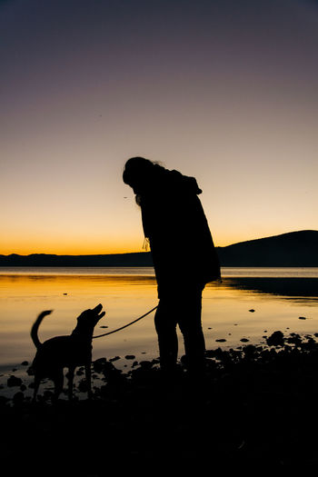 Silhouette woman and dog at lakeshore against clear sky during sunset