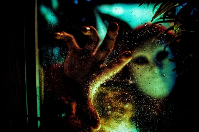 Person wearing mask seen through wet glass window at night