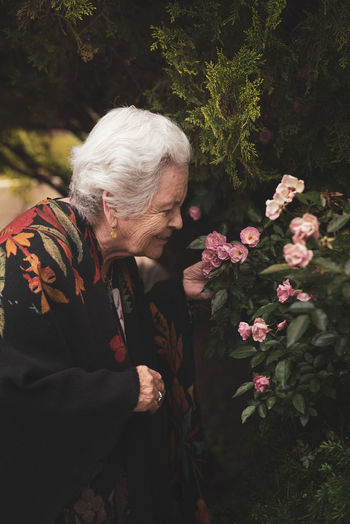 Side view of elderly female standing near blooming bush of pink roses while touching and smelling fresh flowers on summer day