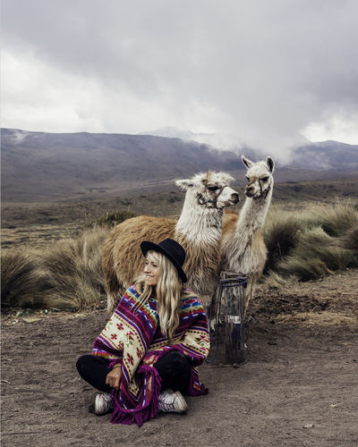 Woman wearing hat sitting by llamas on land