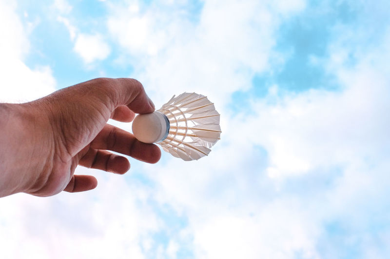 Cropped hand of man holding shuttlecock against cloudy sky