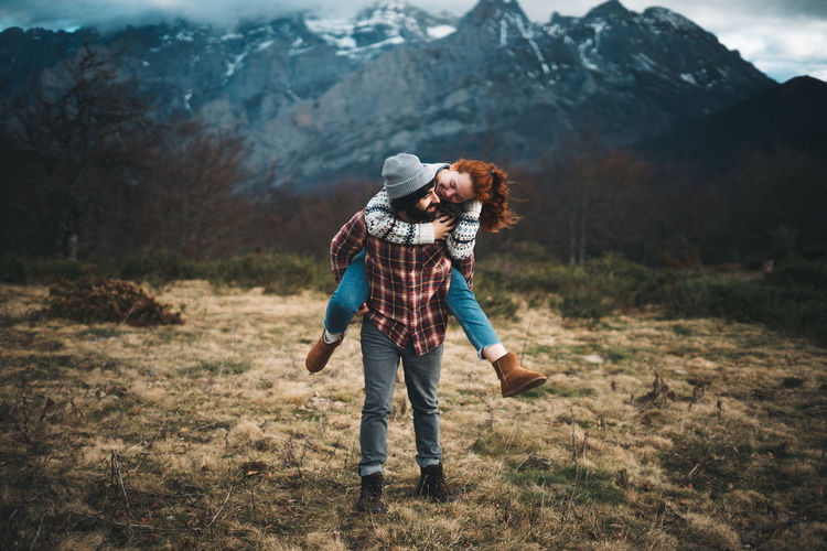 Lovely couple hugging and spending time together piggybacking and traveling in lawn with dry grass near by mountains in cloudy day