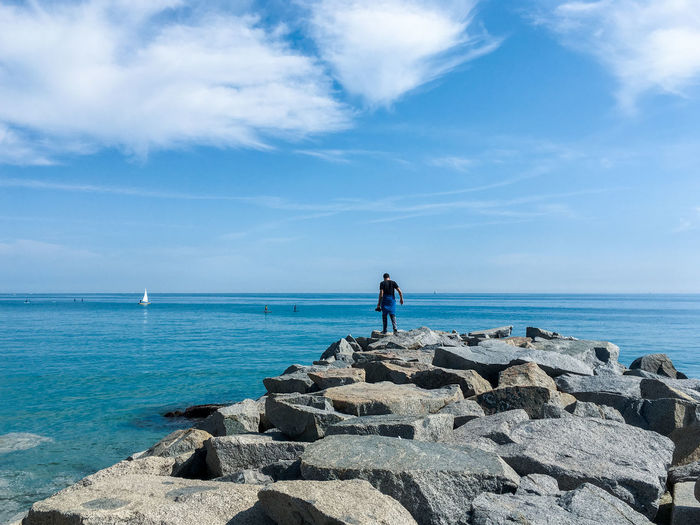 Rear view of man standing on rocks by sea against blue sky