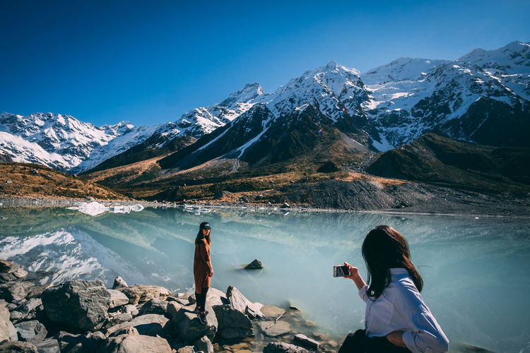 Girls taking photo with her smart phone next lake and snowcapped mountains