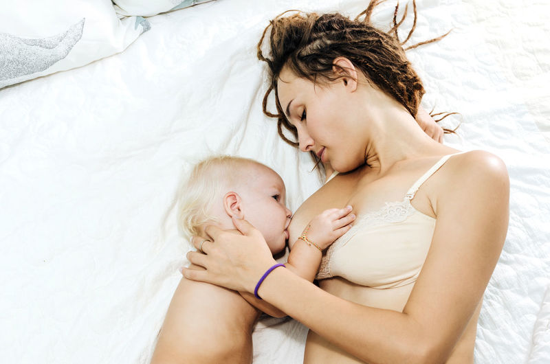 High angle view of mother breastfeeding shirtless daughter on bed at home