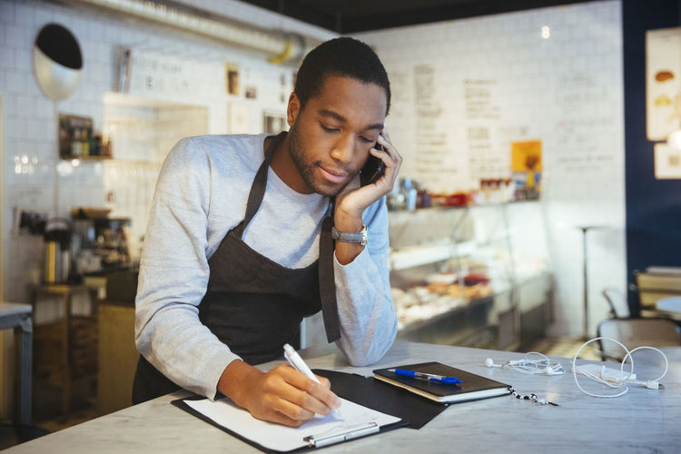 Confident young male employee talking through smart phone while writing on clipboard at delicatessen
