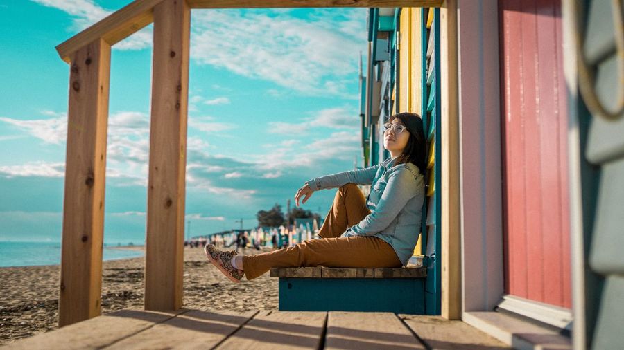 Woman sitting on bench at beach against sky