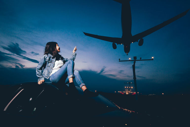 Smiling woman sitting on car with airplane landing at airport against blue sky