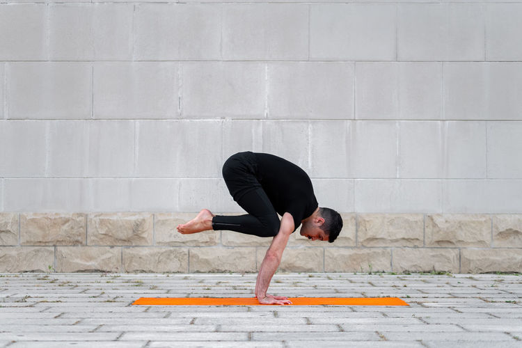 Latin man practicing yoga in a city, standing in crane pose on orange mat