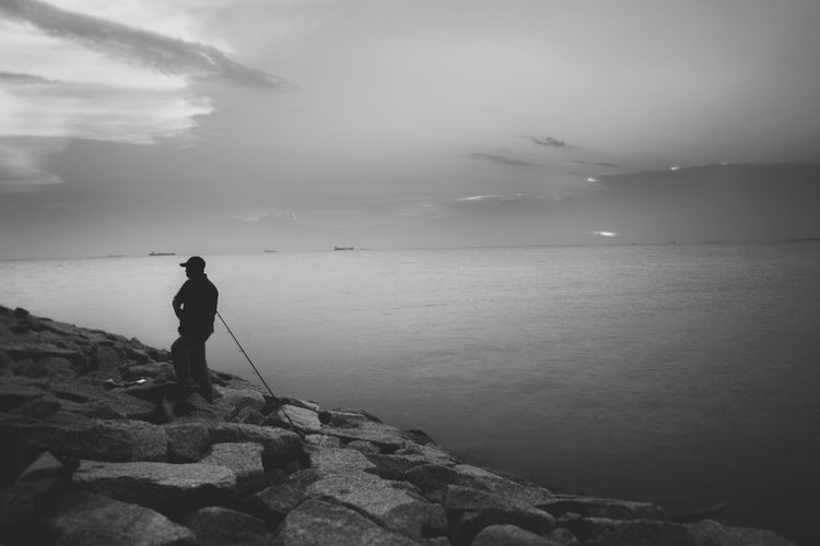 Fisherman standing on rock against sea during sunset