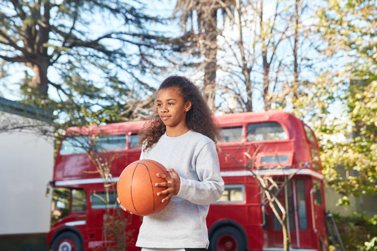 Girl looking away while standing with basketball against trees