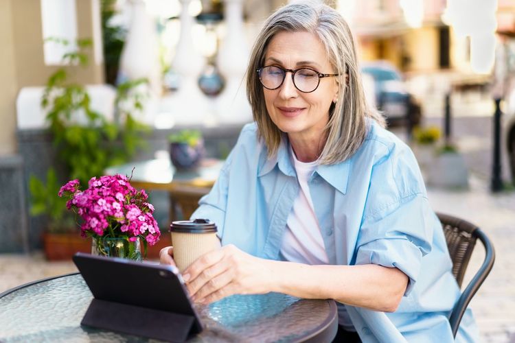 Silver-haired mature european woman sitting at cafe, smiling with joy as she looks at her tablet pc