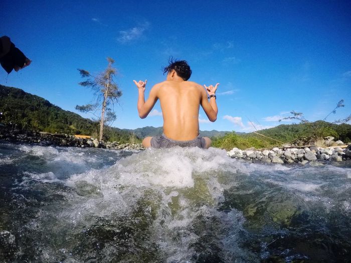 Rear view of man sitting on stream while doing shaka sign against sky