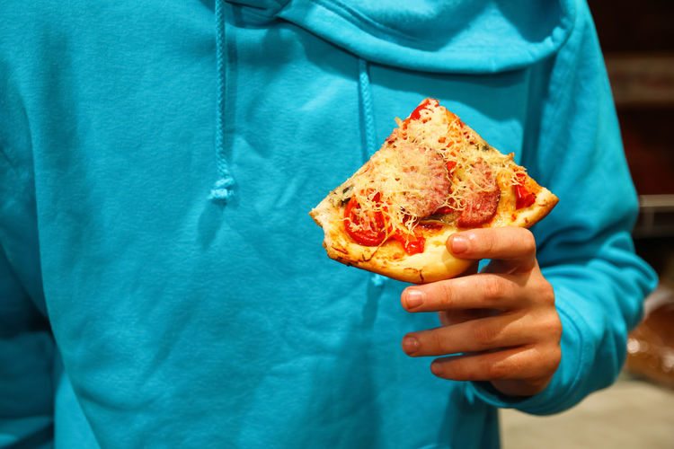 Male hand holding pizza slice. slice of pepperoni pizza in hand isolated on blue. concept 
