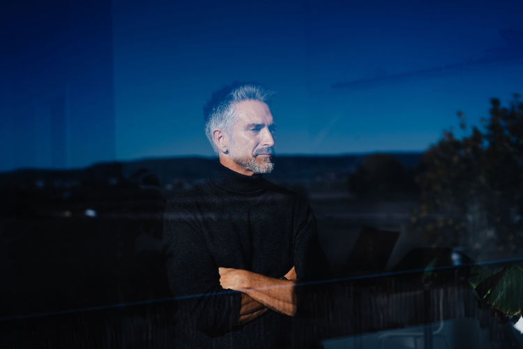 Gray haired senior male standing with crossed arms behind window reflecting landscape under cloudless sky in evening