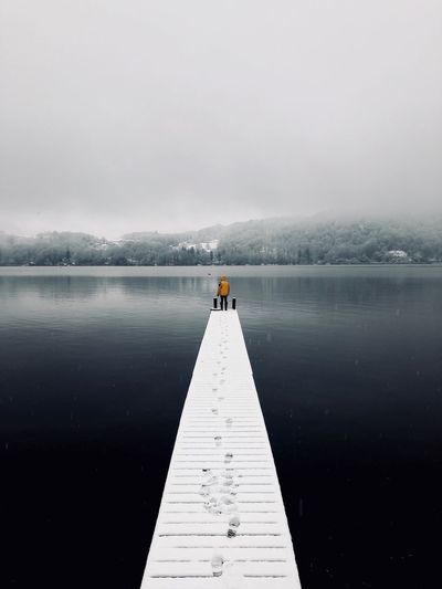 Rear view of person standing on pier over lake against sky during winter