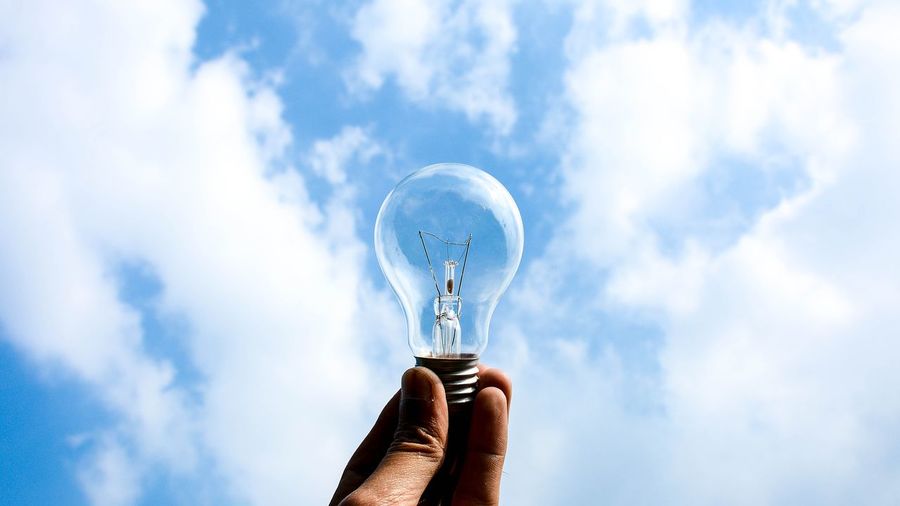 Low angle view of person hand holding light bulb against sky