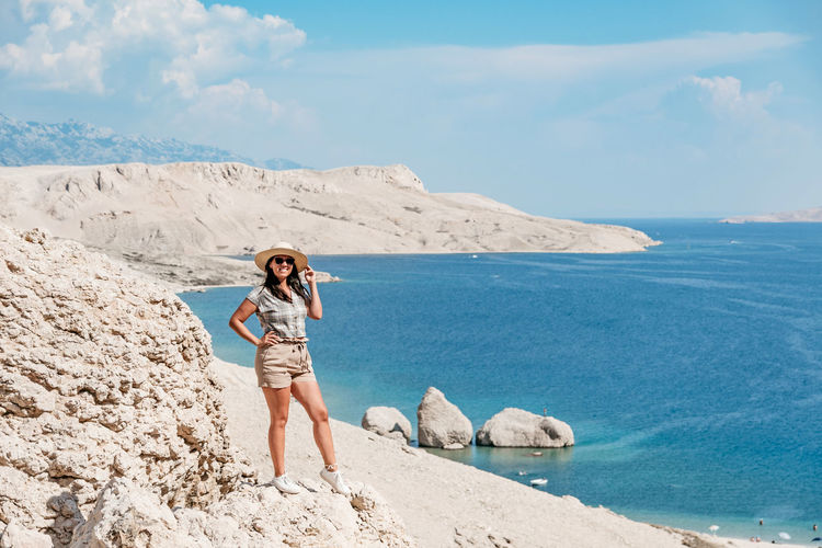 Front view of woman in summer clothes and sun hat standing on cliff overlooking spectacular beach.