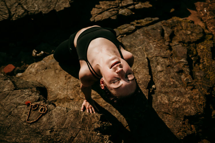 From above of calm female practicing yoga in ustrasana with closed eyes on rocky shore in summer