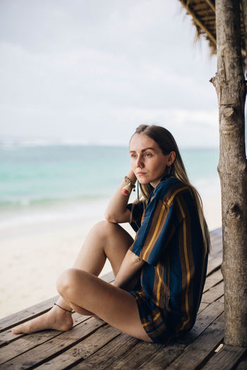 Young woman sitting on shore at beach against sky
