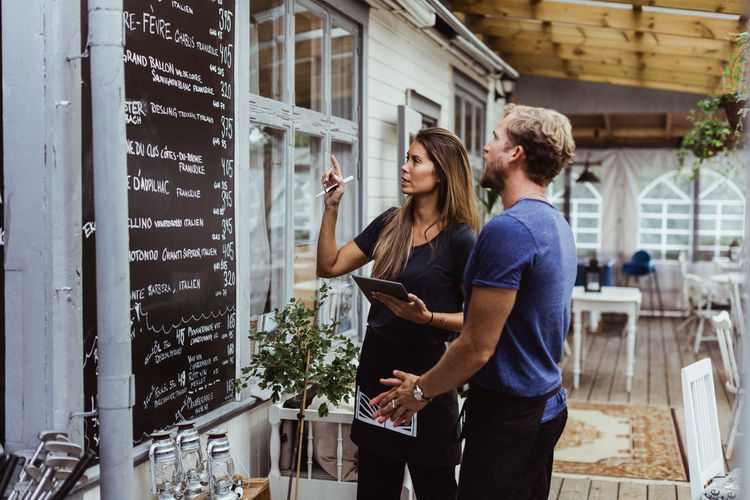 Female owner with digital tablet standing with coworker against menu wall in restaurant