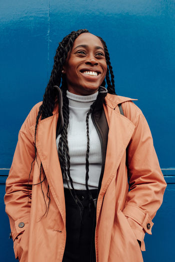 Smiling young woman wearing overcoat while standing against blue wall