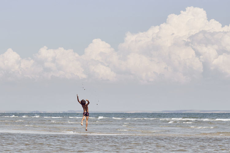 Rear view of shirtless boy jumping in sea against cloudy sky