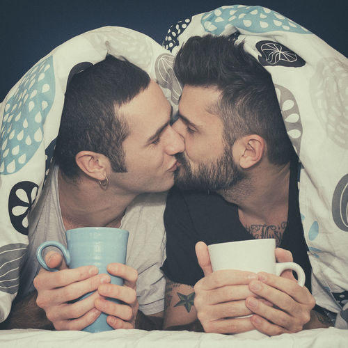 Close-up of homosexual couple holding mugs while kissing under blanket