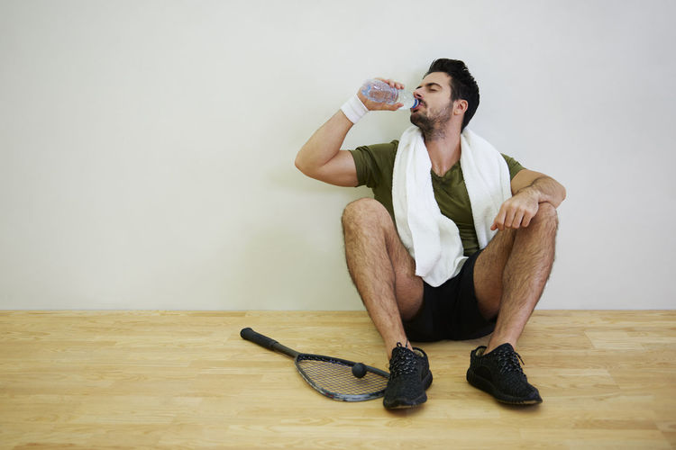 Full length of man drinking water while sitting on floor against wall