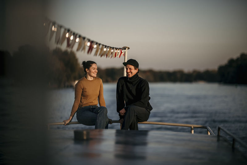 Smiling woman with friend sitting on railing of boat at dusk