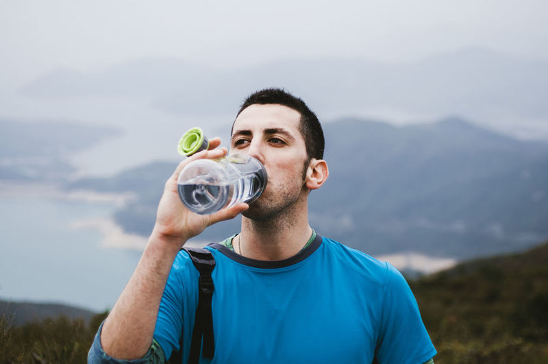 Male hiker drinking water while standing on mountain against sky