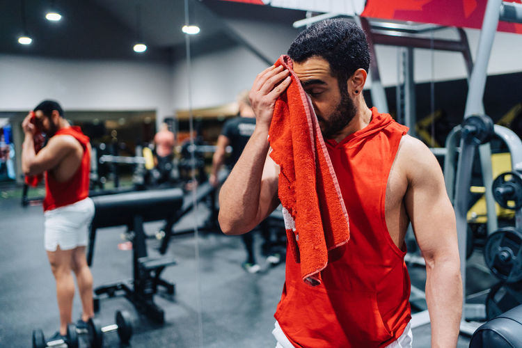 Side view of young man standing in gym cleaning his forehead from sweat after gym exercises 