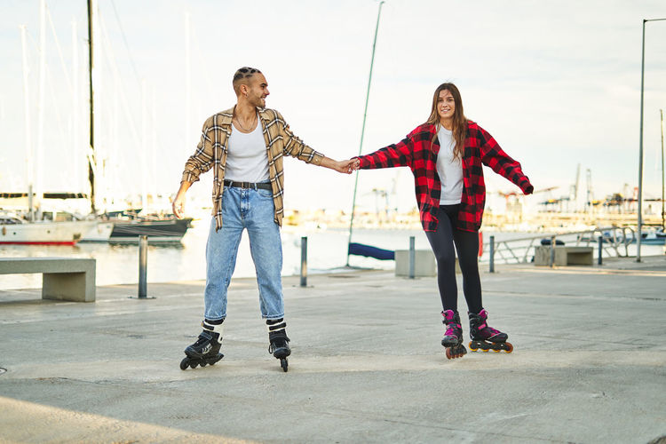 Full body of positive young friends holding hands on roller skates enjoying pleasant free time together on seafront at sunset