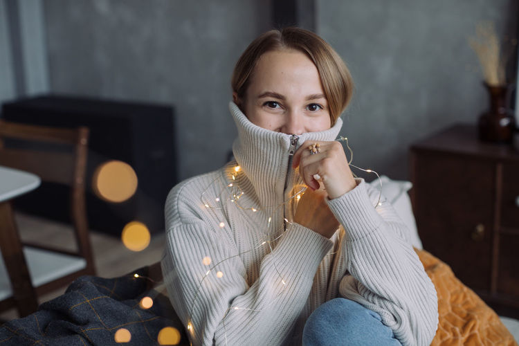 Portrait of woman wearing sweater with illuminated christmas lights at home
