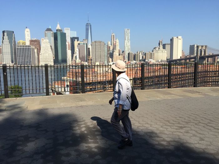 Full length of man walking on footpath by one world trade center and city against clear blue sky