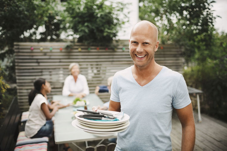 Portrait of happy man holding plates with family sitting at outdoor table in yard
