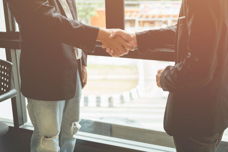 Midsection of businessmen shaking hands while standing by window in office