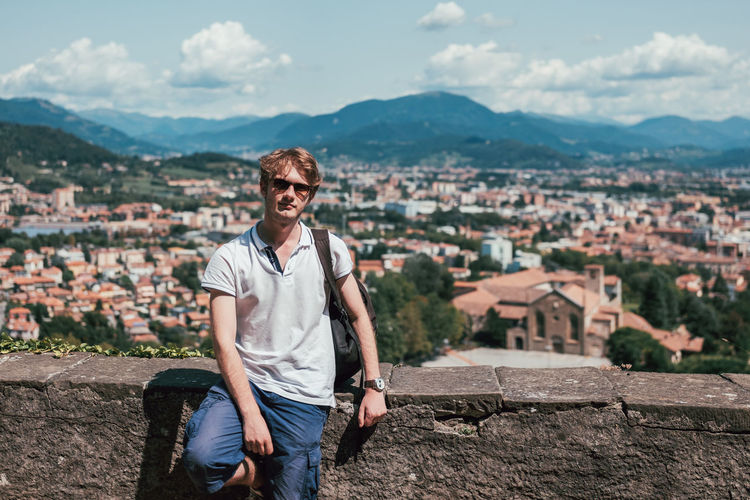 Portrait of young man wearing sunglasses standing by retaining wall against townscape