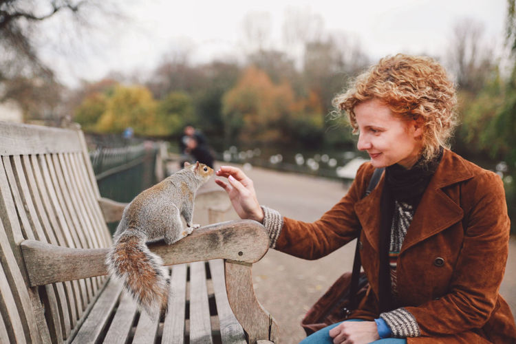 Smiling woman playing with squirrel on bench in park