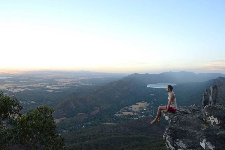 Side view of man sitting on cliff at grampians national park against sky during sunset