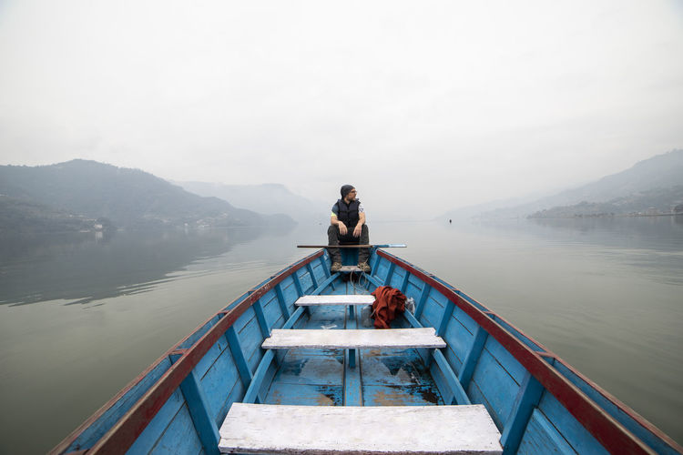 Man sitting on lake by mountains against sky