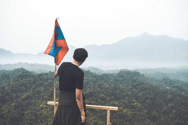 Rear view of man by flag standing on mountain against sky