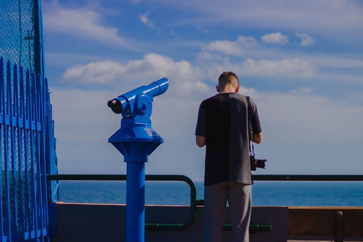 Rear view of man standing by coin-operated binoculars at railing against sea at observation point